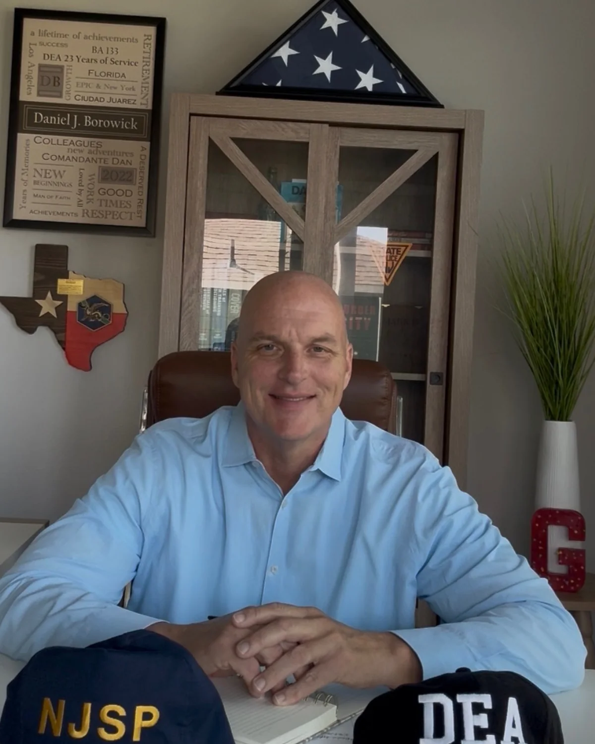 Daniel Borowick at his desk with a notebook, his DEA and New Jersey State Police caps in front of him, and his DEA retirement plaque and folded flag behind